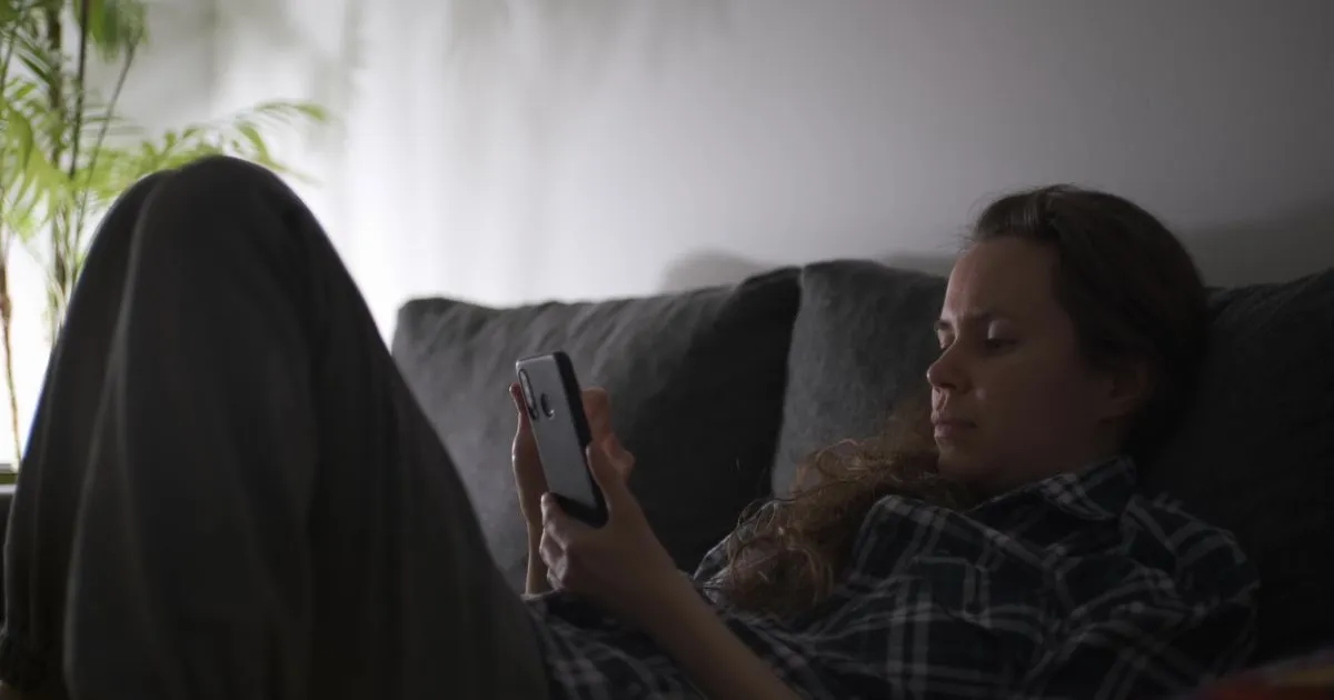 Mulher sentada no sofá da sala à noite, com o celular na mão e a luz baixa do abajur, olhando para a tela com expressão de hesitação antes de enviar uma mensagem