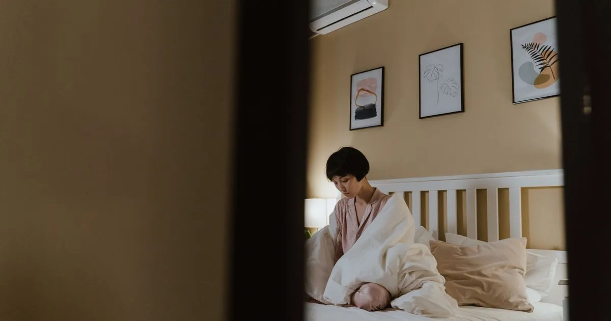 Mulher sentada sozinha na beira da cama de um quarto arrumado, com o olhar baixo e distante, na luz quente do abajur no fim do dia, vista através do vão da porta
