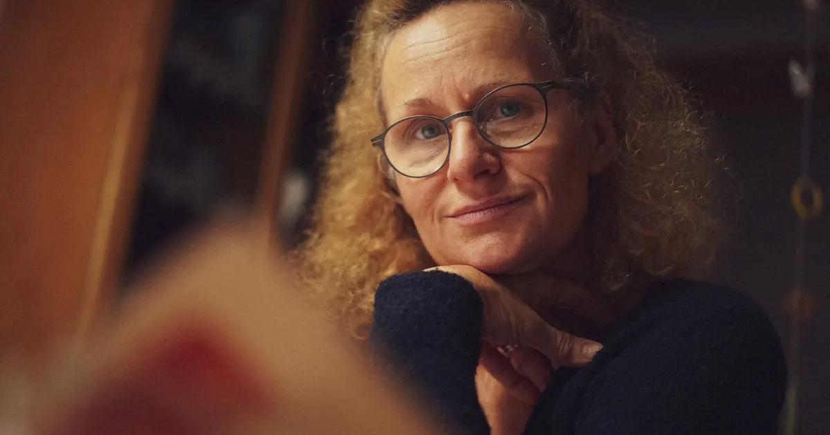 Mulher madura, de óculos, em momento reflexivo sob luz quente, apoiando o rosto na mão
