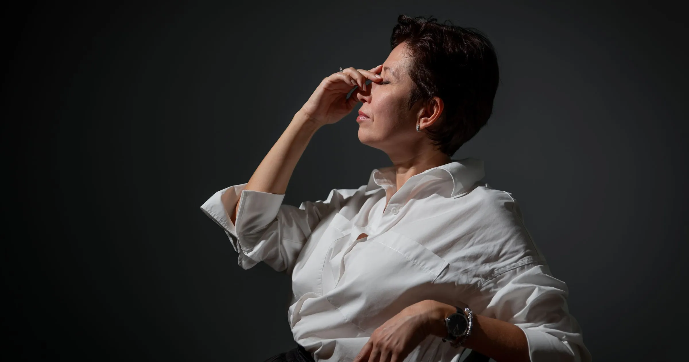 Mulher madura de camisa branca, olhos fechados e mão no rosto, num gesto de cansaço sereno e digno