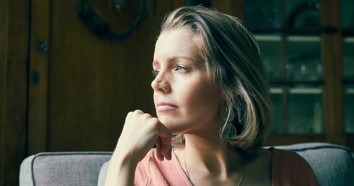 Mulher de meia-idade sentada em casa, sob luz natural quente, olhando para longe com expressão pensativa e a mão apoiada no queixo