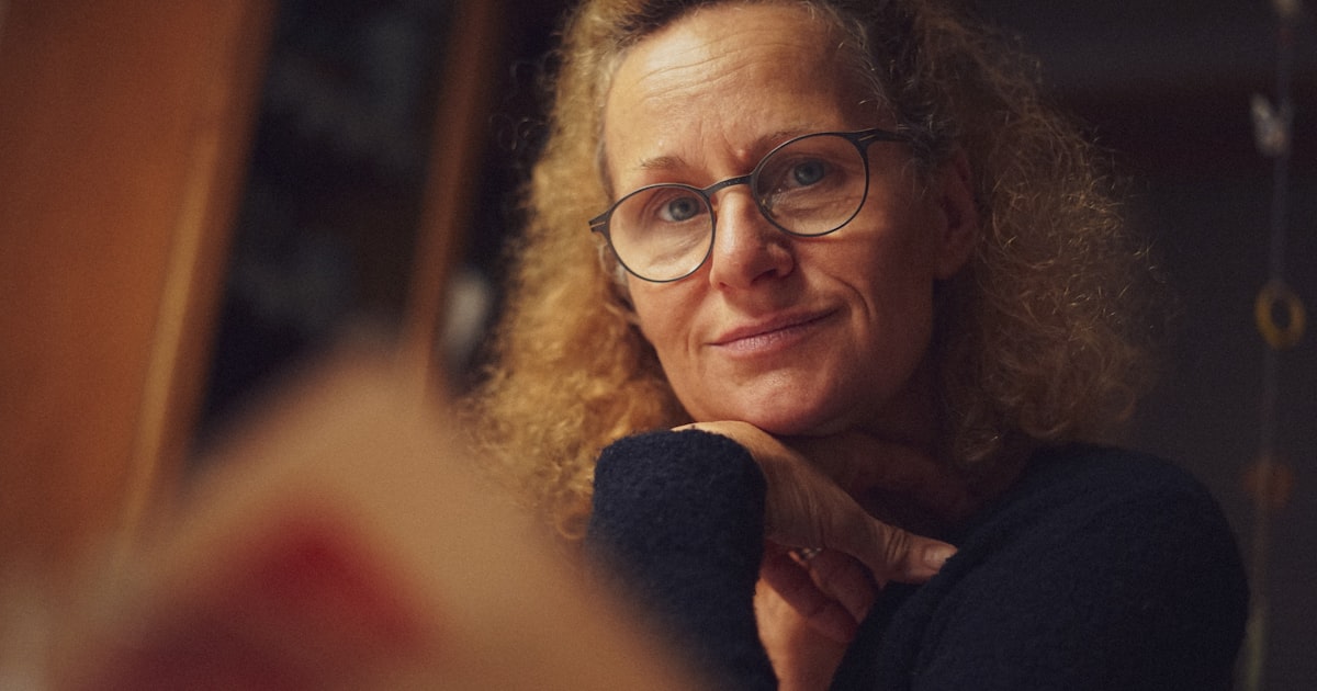 Mulher madura, de óculos, em momento reflexivo sob luz quente, apoiando o rosto na mão