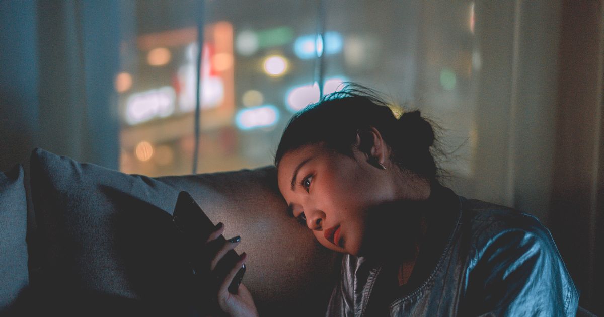Mulher recostada no sofá da sala à noite, com as luzes da cidade desfocadas na janela ao fundo, olhando a tela do celular que ilumina o seu rosto