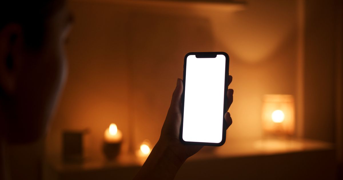 Mão de mulher segurando o celular com a tela acesa no escuro do quarto, iluminada apenas por pequenas luzes quentes ao fundo