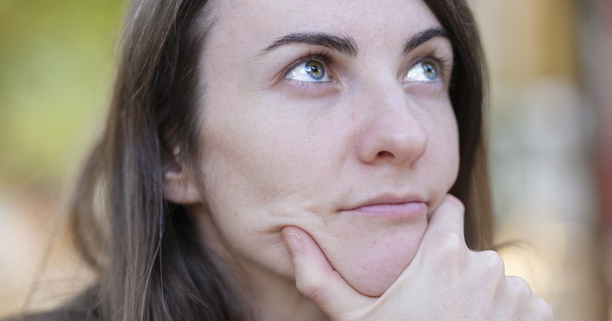 Mulher de meia-idade em pausa reflexiva, mão no queixo e olhar atento, sob luz natural quente