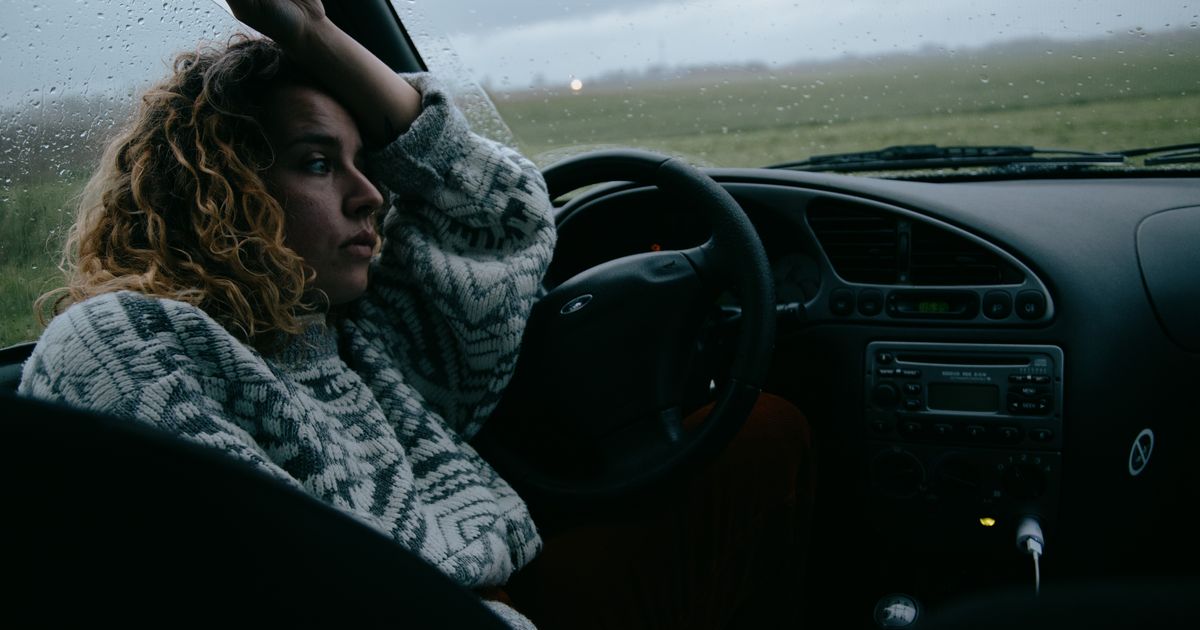 Mulher de cabelos cacheados sentada ao volante de um carro parado, em um dia cinzento, com o braço apoiado junto à janela e o olhar perdido através do para-brisa molhado de chuva
