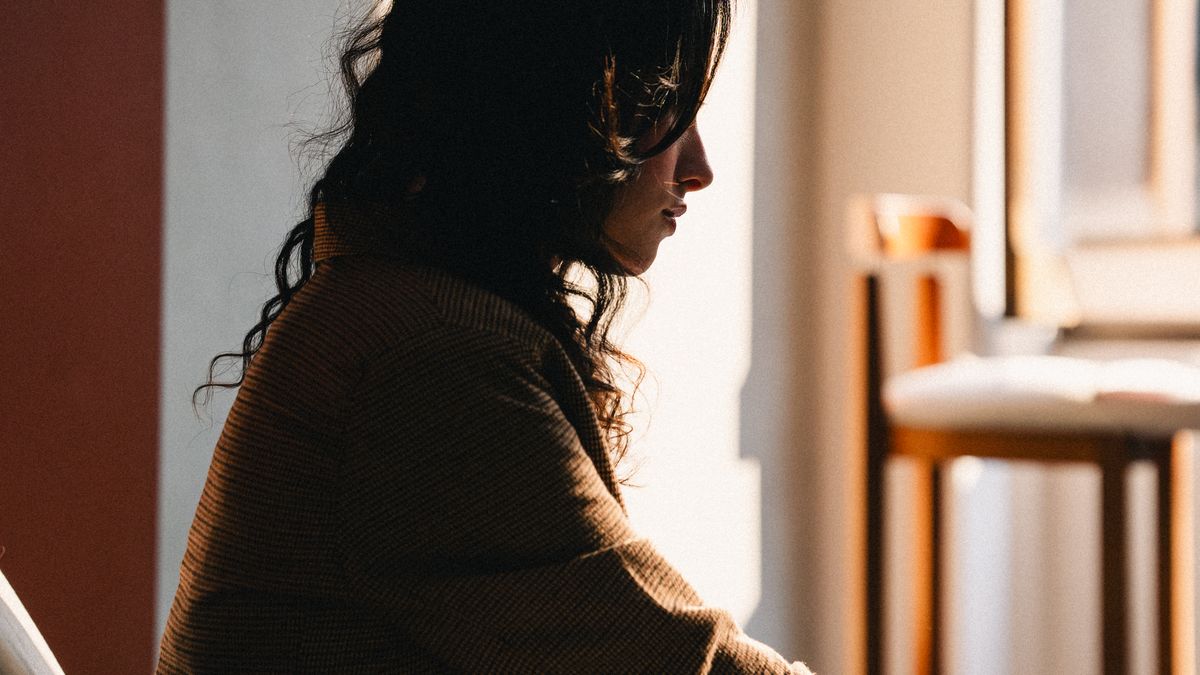 Mulher de perfil sentada num canto silencioso da casa, banhada pela luz quente do fim da tarde, num momento de pausa em que finalmente escuta o que está sentindo
