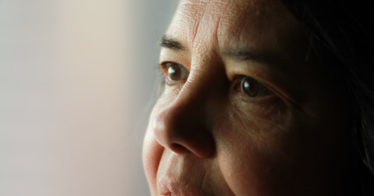 Mulher madura em close de perfil, olhar reflexivo voltado para a luz suave da janela, em tom quente e editorial
