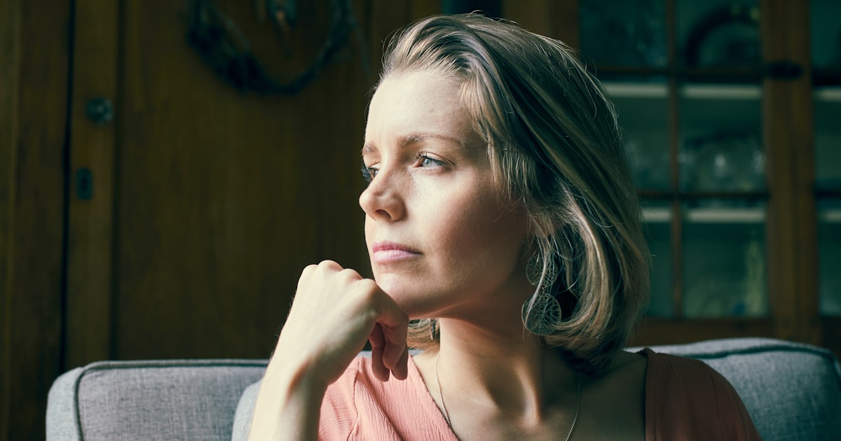 Mulher de meia-idade sentada em casa, sob luz natural quente, olhando para longe com expressão pensativa e a mão apoiada no queixo