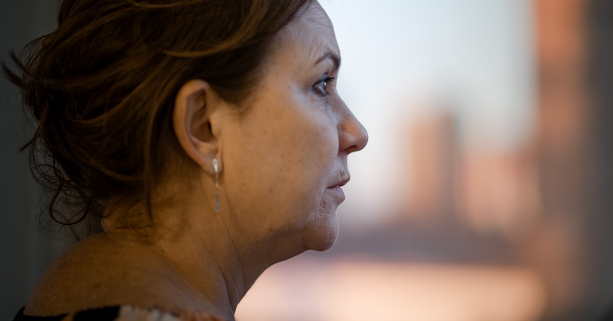 Mulher por volta dos 50 anos de perfil, com o olhar voltado para a luz quente do fim de tarde que entra pela janela, num momento de reflexão silenciosa